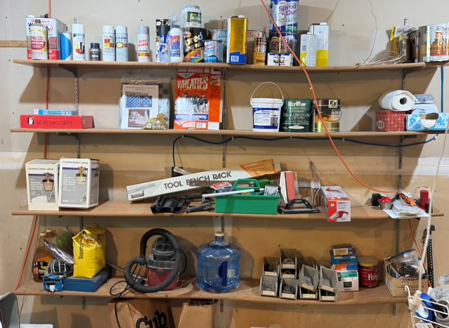 Contents of wooden shelves, including- various paints- gloss trim- Wheaties box- board games- decorative outdoor lighting- tools- vacuum- mini shop back